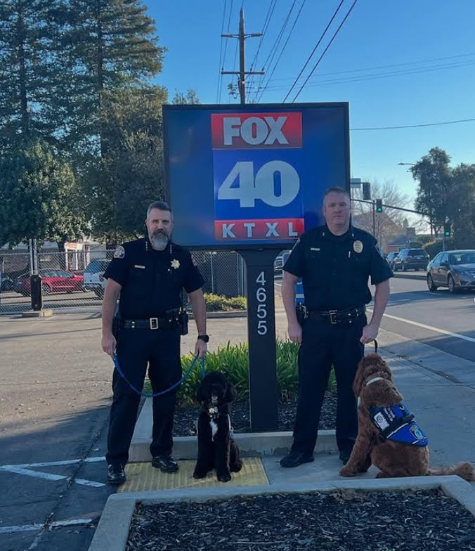 Two uniformed officers with service dogs beside a FOX 40 KTXL news station sign